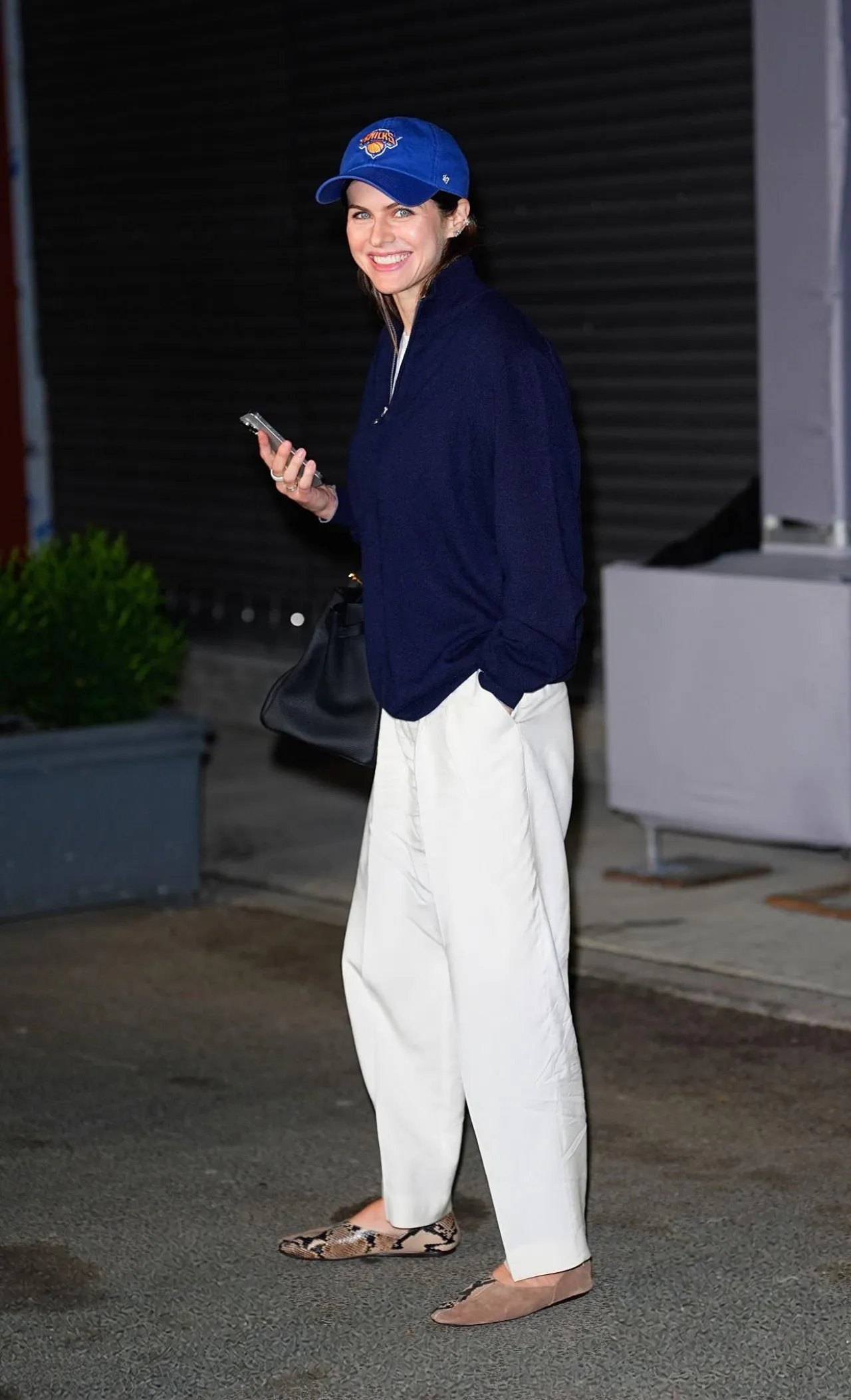 Alexandra Daddario walking in a navy cap and white pants at the US Open