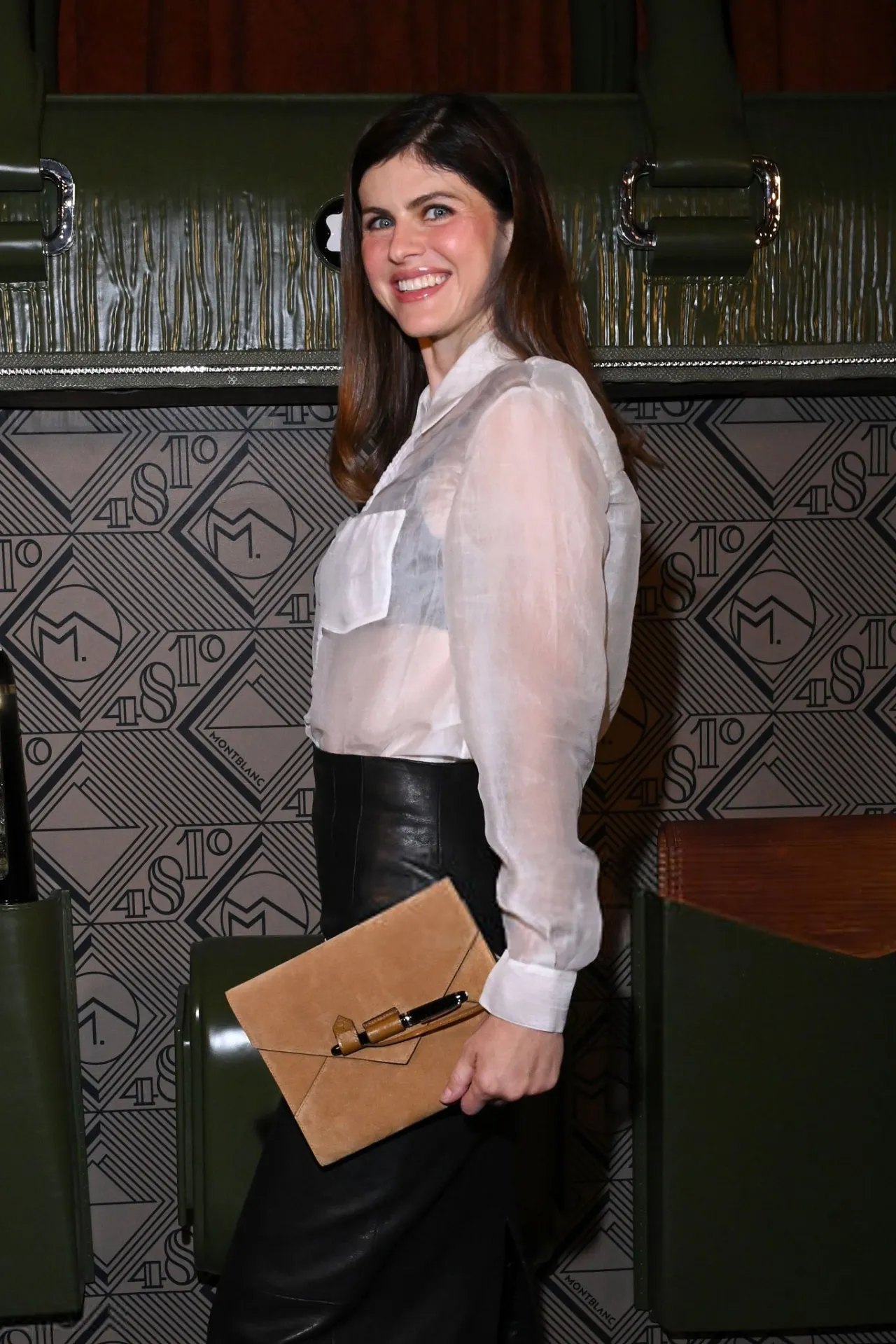 Alexandra Daddario holding a tan clutch in a sheer shirt and leather skirt at Montblanc