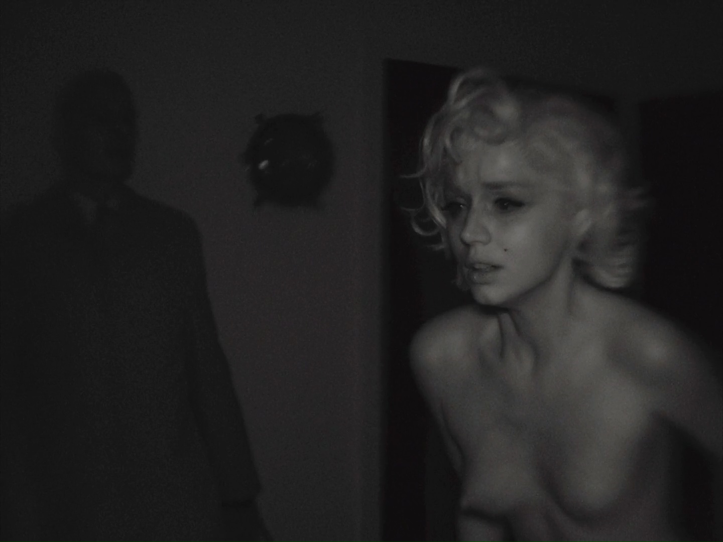 Blonde, her face and bare chest, a man in the dark behind her