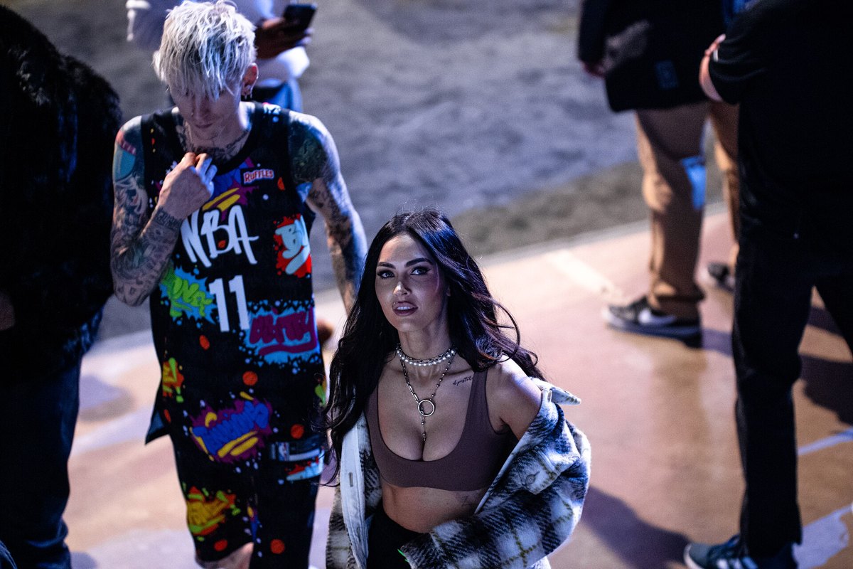 Megan Fox in a brown crop top and plaid coat walking with Machine Gun Kelly in a graffiti NBA jersey, February 2022
