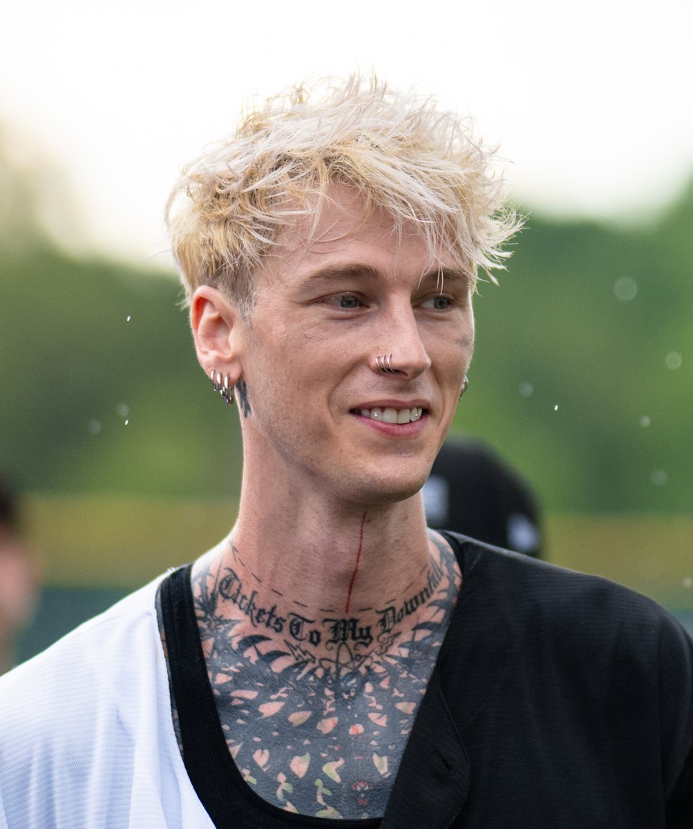 Machine Gun Kelly with bleached hair and neck tattoos smiling at a charity softball game, 2023