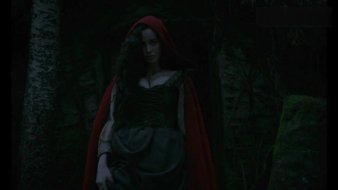Sarah Stephens in a red hood and laced bodice at the doorway of a hut in The Witch