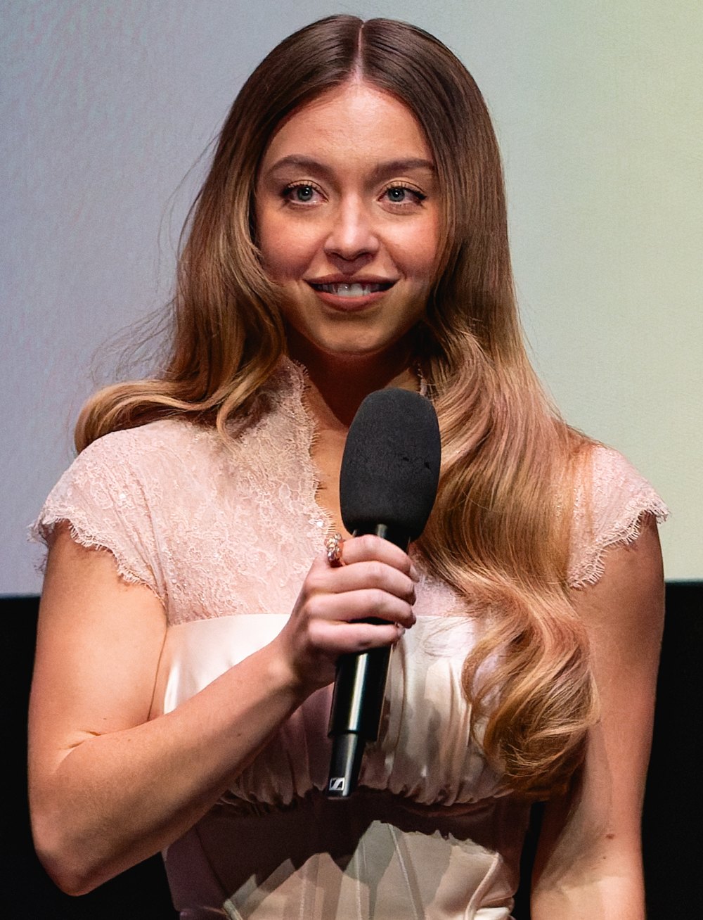 Sydney Sweeney smiling with a microphone in a blush lace-sleeve dress on stage at the 2025 BFI London Film Festival
